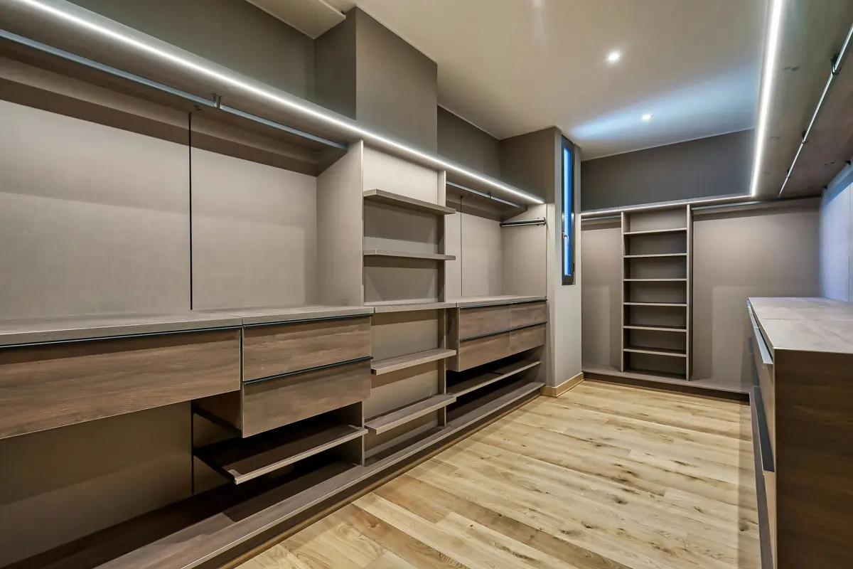 Closet planejado moderno e espaçoso com acabamento em madeira clara e iluminação LED embutida, incluindo gavetas, prateleiras, nichos e cabideiros organizados de forma funcional.